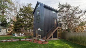 Une maison moderne à deux étages, dotée d’une façade sombre, d’un porche avec un espace salon extérieur et d’un escalier menant à l’étage supérieur.