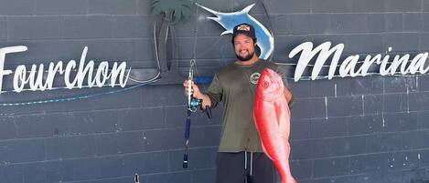 Un homme tenant un gros poisson devant un mur sur lequel on peut lire le texte « Fourchon Marina ».