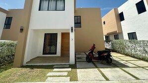 A modern two-story residential building with a flat roof, a central entrance with a wooden door, and a small garden area with a paved walkway.