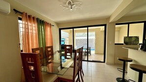 A dining area with a glass table and wooden chairs, a ceiling fan, and a sliding glass door leading to a balcony.