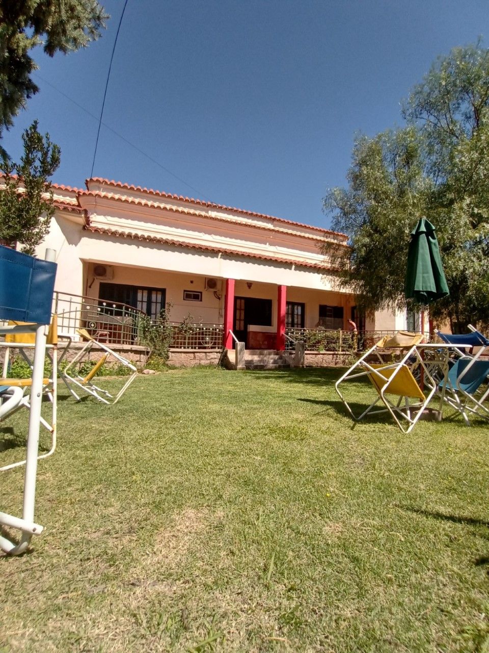 Una casa con techo de tejas, puerta roja y un pequeño porche. Hay sillas de exterior y un paraguas verde.