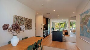 A modern kitchen with a central island, a dining area with a wooden table and chairs, and a view of the outdoors through large windows. - Malthouse Lane City Apartment 4 with Stunning Hill Views in Nelson CBD (Nelson)