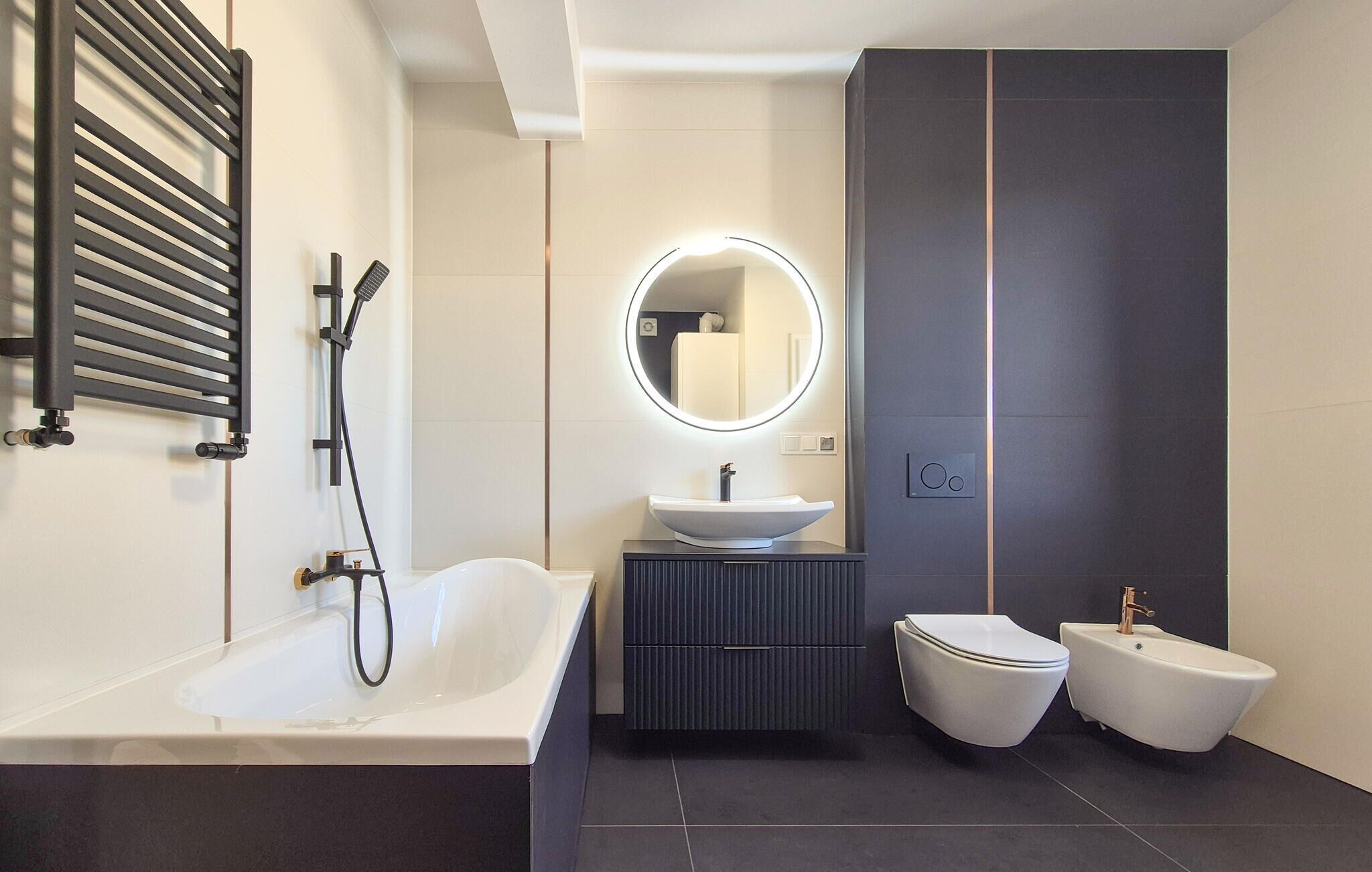 Een moderne badkamer met een vrijstaand bad, een ronde spiegel met verlichting, een wastafel op een zuil en twee bidets.