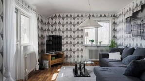 Un salon moderne avec un canapé gris, une table basse en verre et un téléviseur. La pièce est décorée d’un papier peint à motifs et possède une fenêtre avec des rideaux.