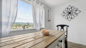 A room with a wooden table, a chair, a window with sheer curtains, and a wall-mounted decorative metal piece.