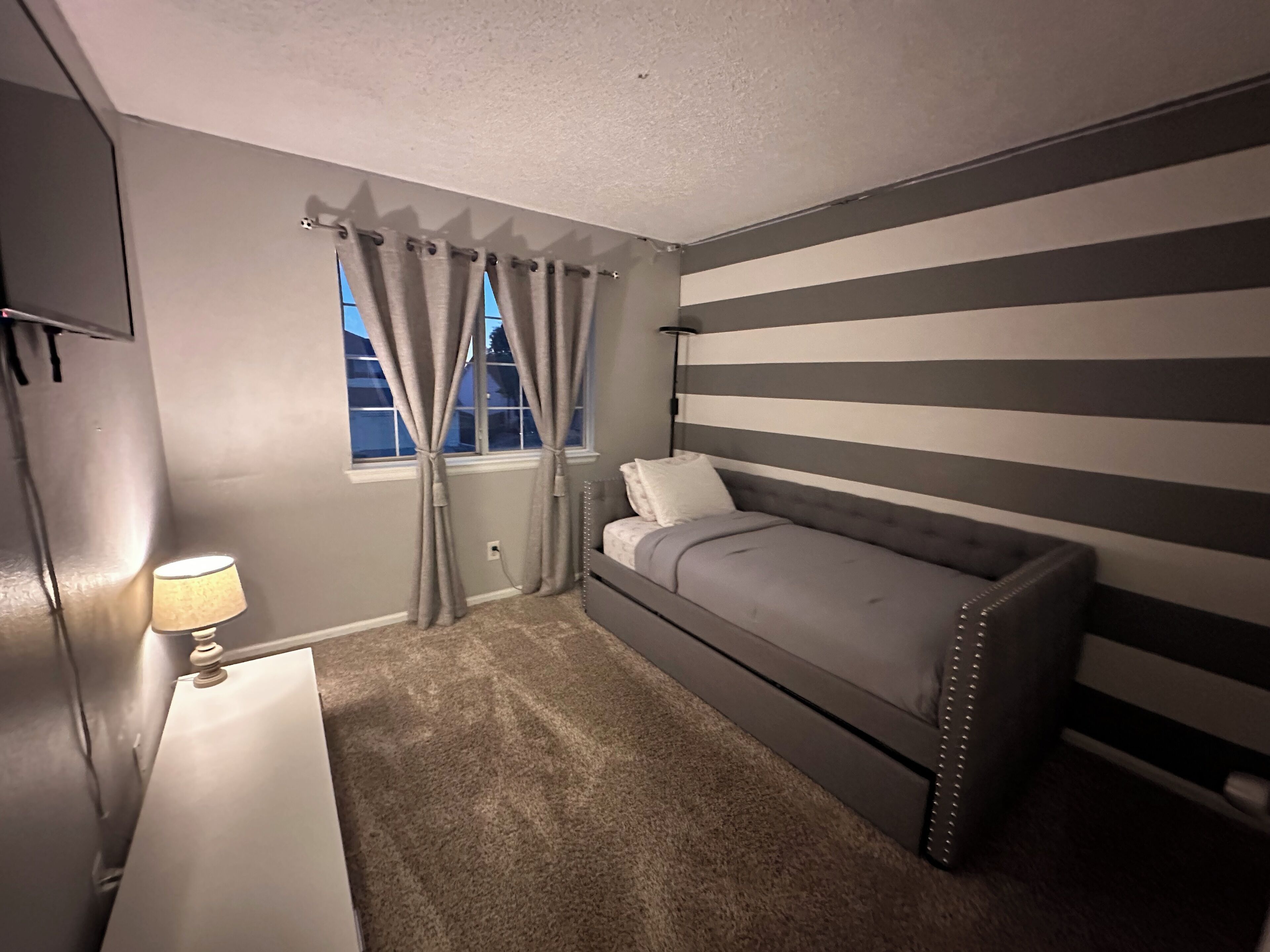 A bedroom with a bed, a nightstand, a lamp, a window with curtains, and a striped wall.