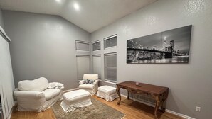A room with a white armchair, a matching ottoman, a small table, and a cityscape painting on the wall. - Charming Broomfield Retreat: Modern Comforts & Prime Location
 (Broomfield)