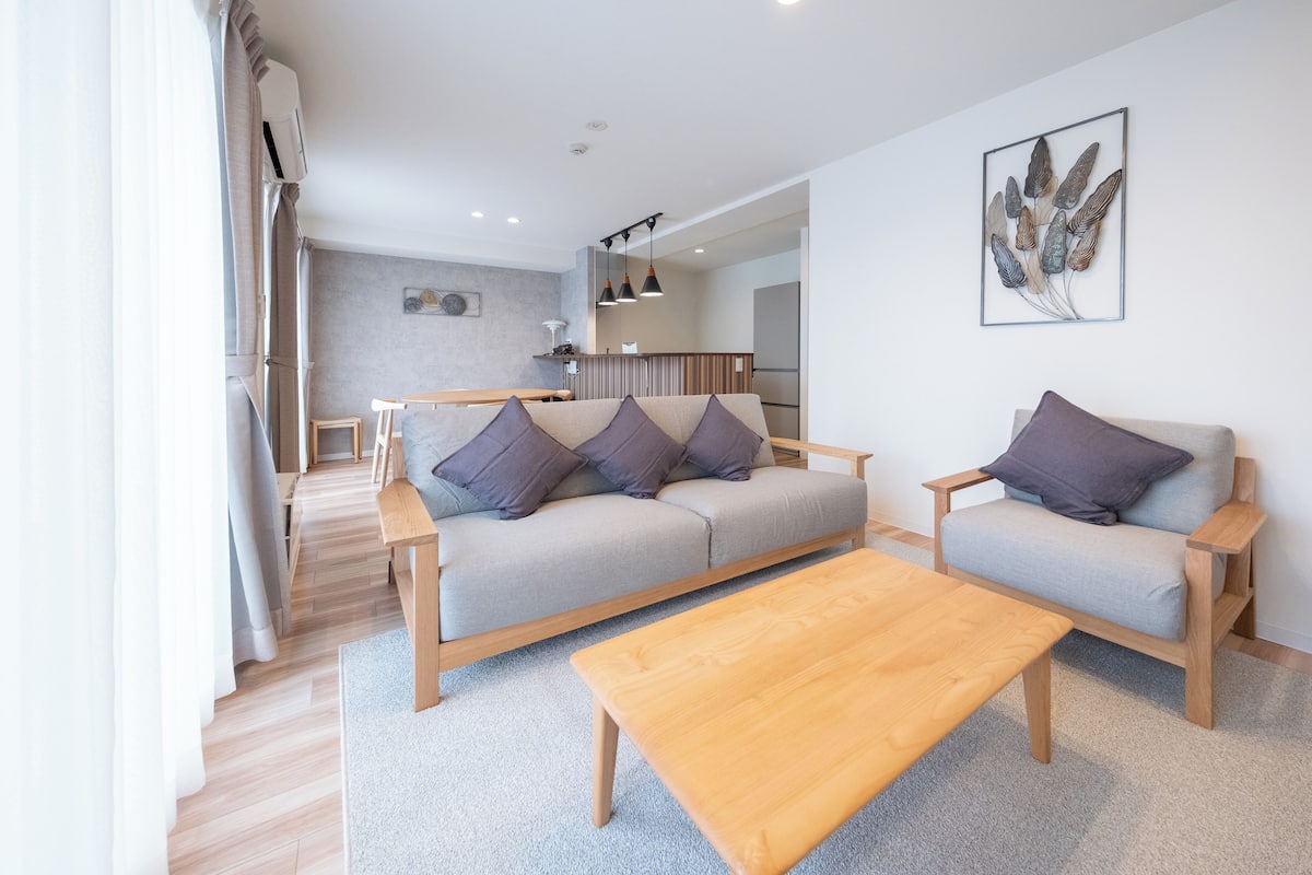 Una sala moderna con un sofá, dos sillones, una mesa de centro y una cocina al fondo.