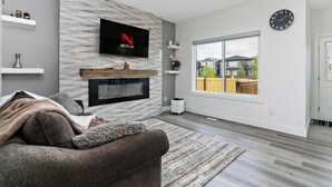 A modern living room with a flat-screen TV, fireplace, and large window. - Luxurious, Cozy, & Modern 4 bedroom Home—Airconditioned + Netflix + Free Parking (Edmonton)