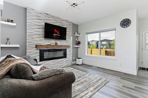 A modern living room with a flat-screen TV, fireplace, and large window. - Luxurious, Cozy, & Modern 4 bedroom Home—Airconditioned + Netflix + Free Parking (Edmonton)
