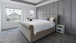 A bedroom with a large bed, a nightstand, a window, and a view of houses outside. - Luxurious, Cozy, & Modern 4 bedroom Home—Airconditioned + Netflix + Free Parking (Edmonton)