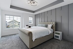 A bedroom with a large bed, a nightstand, a window, and a view of houses outside. - Luxurious, Cozy, & Modern 4 bedroom Home—Airconditioned + Netflix + Free Parking (Edmonton)