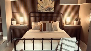 Ein modernes Schlafzimmer mit einem großen Bett, einer Bank, zwei Nachttischen mit Lampen sowie einem an der Wand befestigten Kunstwerk.