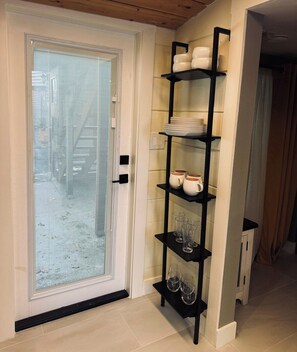 Una porta con vetro acidato, un mobiletto in metallo nero con piatti, bicchieri e calici, e un pavimento piastrellato.