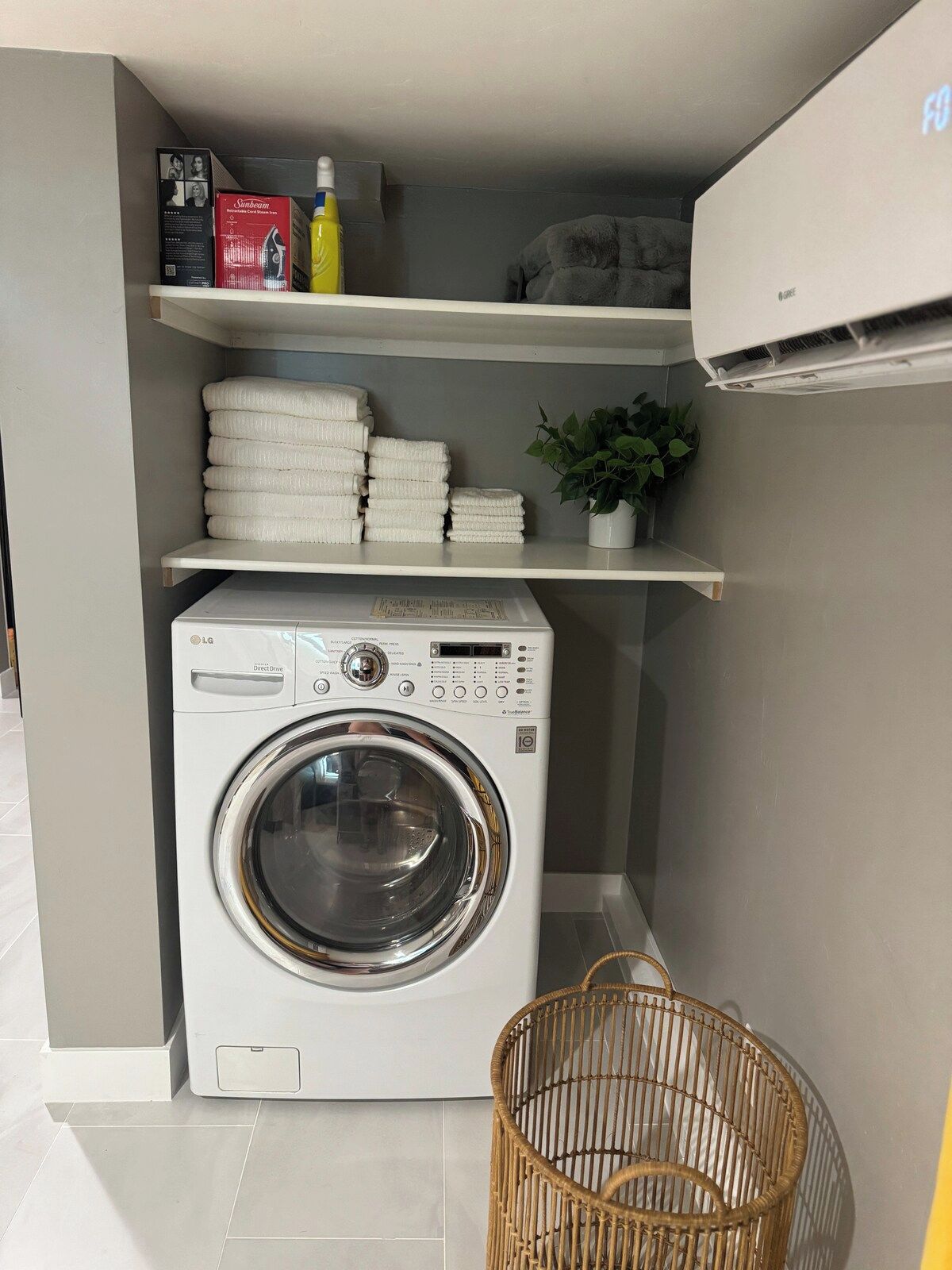 Un espacio de lavandería pequeño con una lavadora de carga frontal, una cesta de mimbre y un estante con toallas dobladas y una planta en maceta.