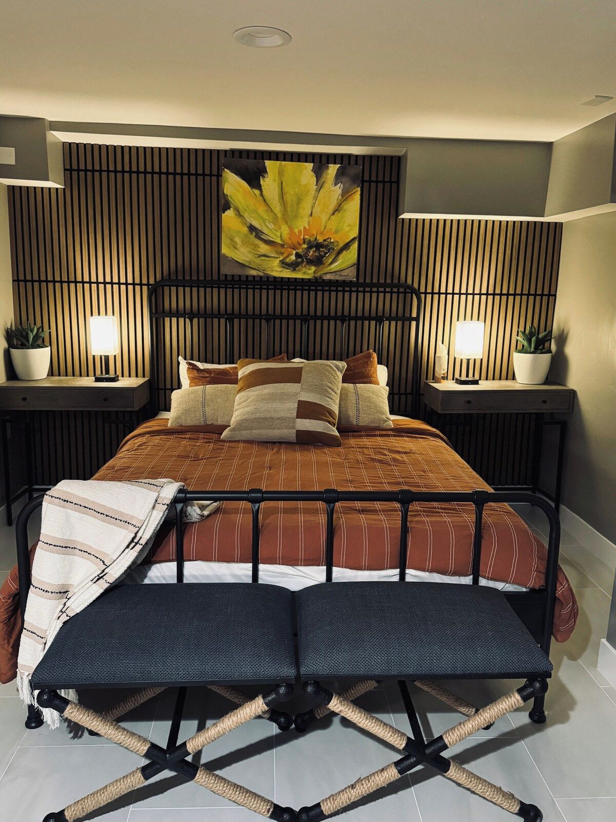 Una cama con cabecera, mesitas de noche con lámparas, una obra de arte en la pared y una planta en maceta.