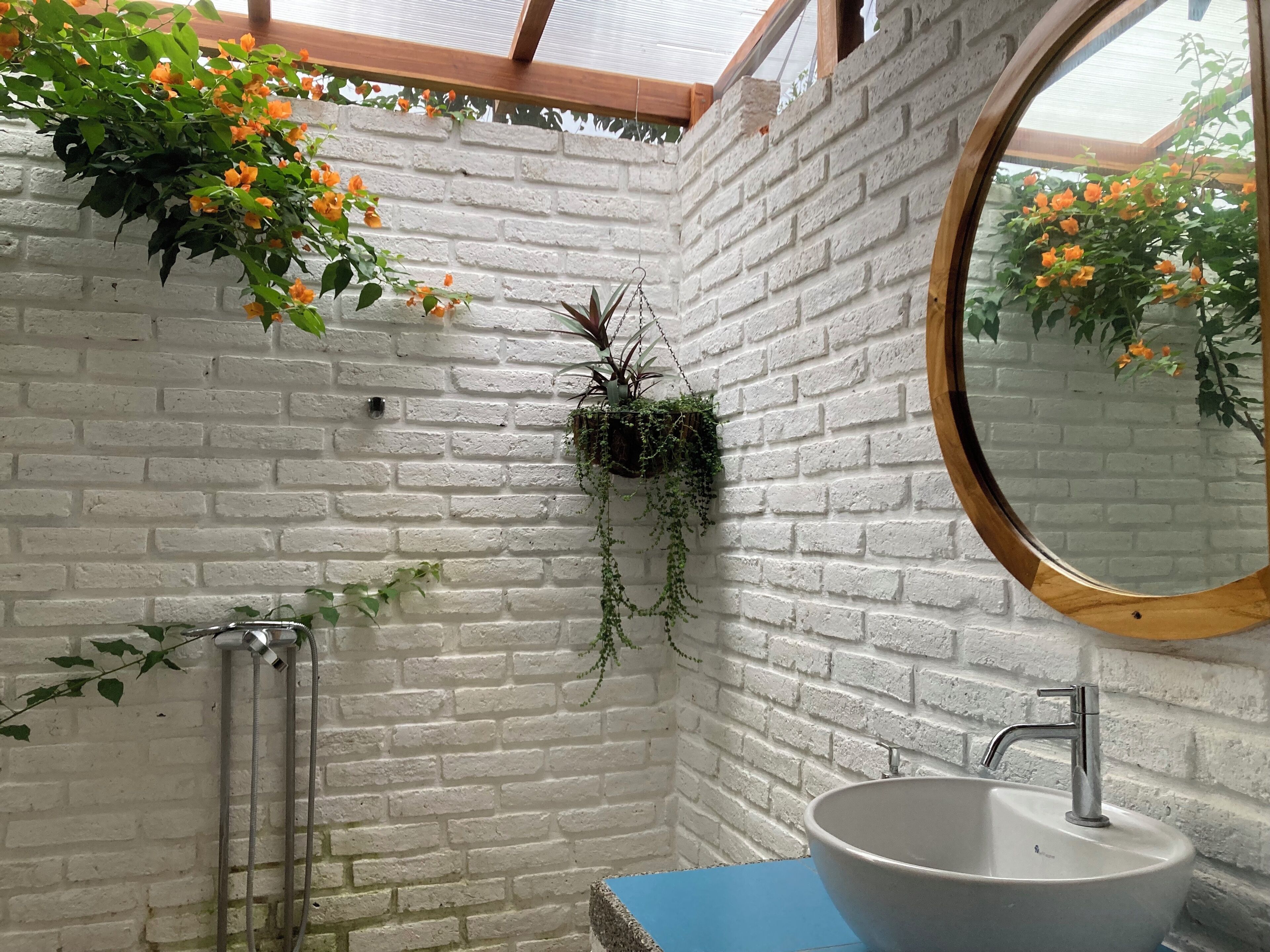 Un baño con pared de ladrillo blanco, espejo redondo, lavamanos y plantas colgantes.