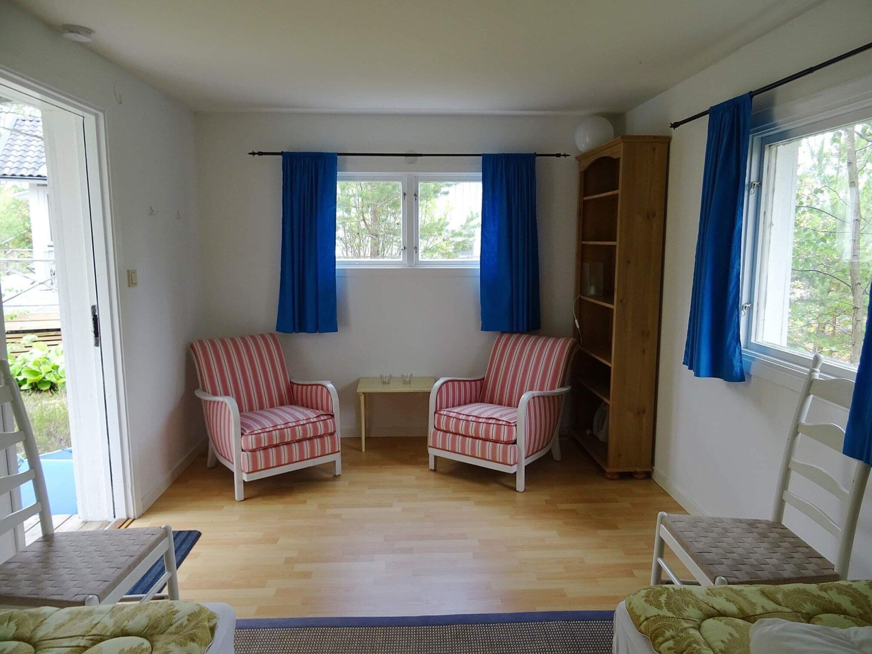 Ein Zimmer mit zwei gestreifte Sesseln, einem kleinen Tisch, einem Bücherregal und einem Fenster mit blauen Vorhängen.