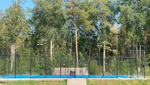 A fenced tennis court with a blue surface, surrounded by trees and a metal fence. - 4 Star Holiday Home in Vaxholm (Vaxholm)