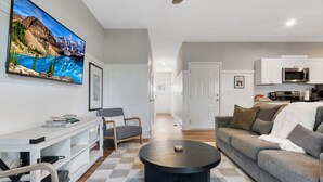 A modern living room with a checkered floor, a grey sofa, a coffee table, and a TV mounted on the wall displaying a mountain landscape. - Ducks Landing Accommodations at Southern Pines Golf Course (Aberdeen)