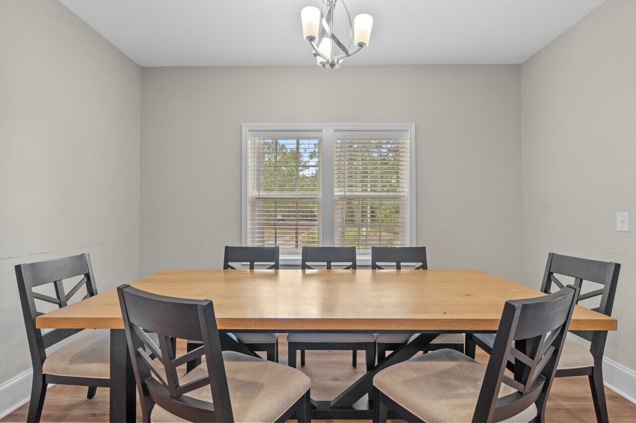 A dining room with a wooden table and six chairs, large windows with blinds, and a chandelier.