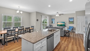 A modern kitchen with granite countertops, white cabinetry, and a dishwasher. There is a dining area with a table and chairs, and a living area with a sofa and a TV. - Ducks Landing Accommodations at Southern Pines Golf Course (Aberdeen)