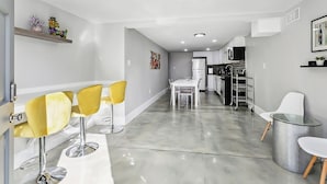 Una cocina moderna con armarios blancos, un refrigerador de acero inoxidable y un microondas. Hay una zona de comedor con una mesa blanca y sillas amarillas.