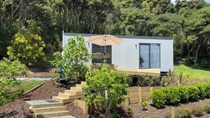 A modern, single-story house with a wooden deck, a staircase leading to a covered patio, and a lush garden. - Te Henga Hideaway  (Te Henga (Bethells Beach))