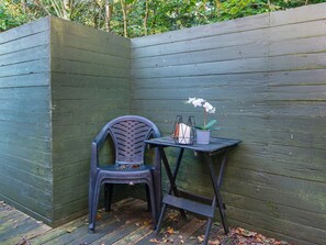 Un rincón con una valla de madera verde oscuro, una silla de plástico, una mesita con una orquídea en maceta y un portavelas.