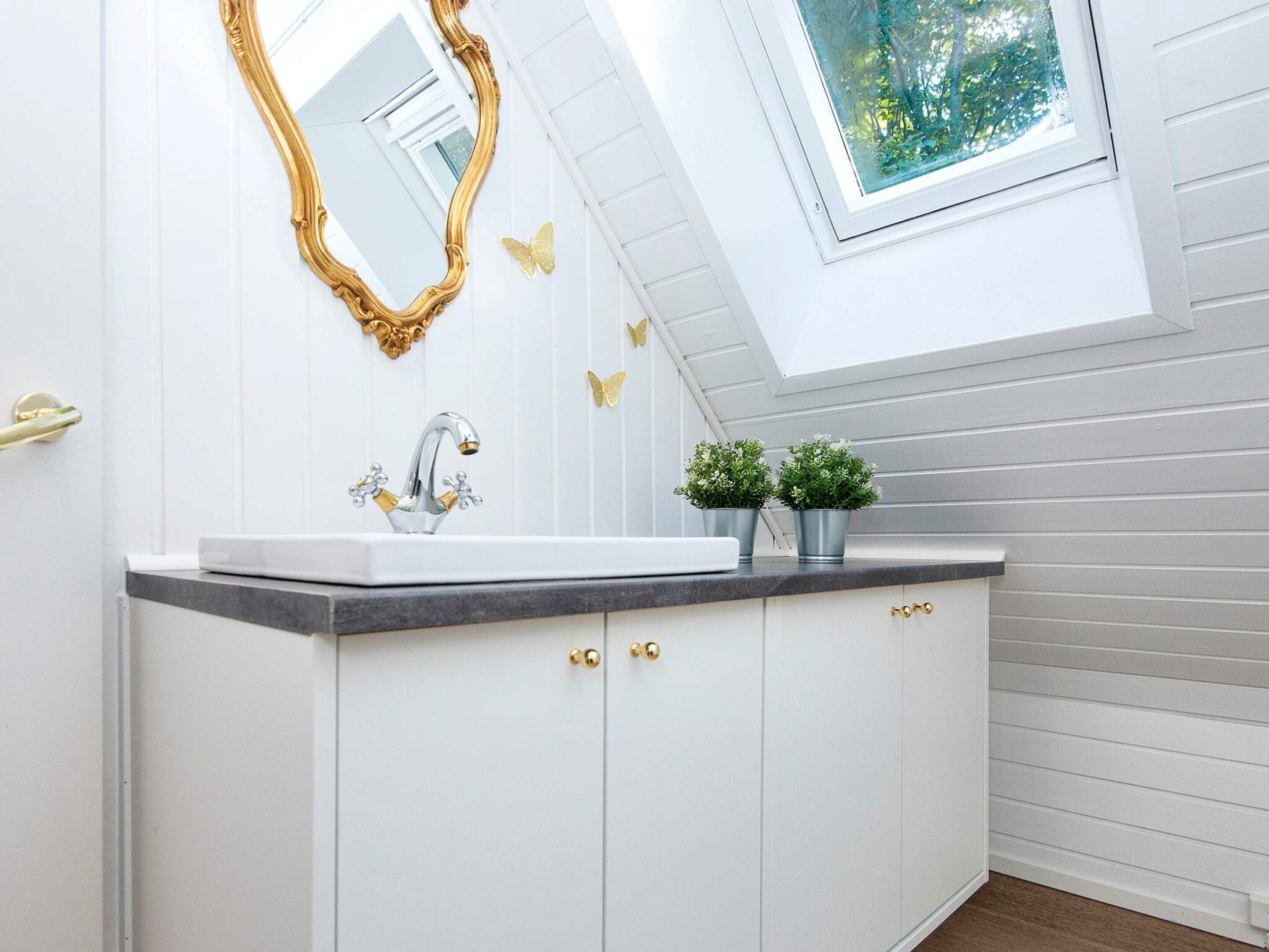A bathroom with a skylight, a white vanity with gold handles, a gold-framed mirror, and two small potted plants.