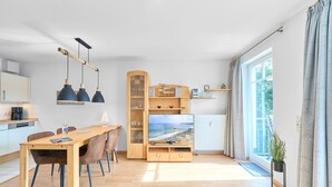 A modern living room with a wooden dining table, chairs, a TV, and a bookshelf. - Residence Johannsens B 12 - Residence Johannsens (Timmendorfer Strand)