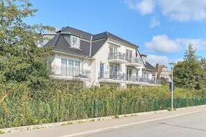 A multi-story residential building with a balcony on the second floor, a sloped roof, and a fence in front.