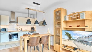A modern kitchen with a dining area, wooden cabinets, and a TV displaying a coastal scene. - Residence Johannsens B 12 - Residence Johannsens (Timmendorfer Strand)