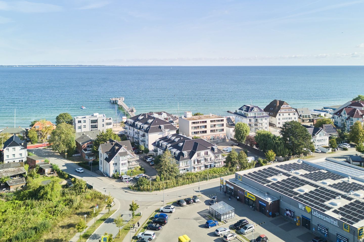 Een kustwoongebied met een mix van eengezins- en meergezinswoningen, een pier die in zee uitloopt, en een bedrijfsgebouw met zonnepanelen.