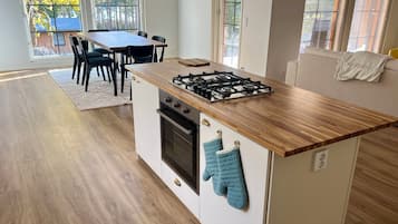 Una cocina moderna con encimera de madera, estufa a gas y horno empotrado. Hay una mesa de comedor con sillas y vistas a árboles a través de ventanas amplias.