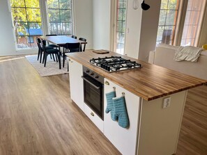 Una cucina moderna con un piano di lavoro in legno, un fornello a gas e forno integrato. C'è un tavolo da pranzo con sedie e vista sugli alberi attraverso ampie finestre.