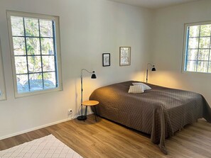 Un dormitorio con una cama grande, dos lámparas de noche y una mesita pequeña. Hay dos ventanas con persianas y un cuadro en la pared.
