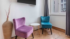 Une pièce dotée d’une grande fenêtre, d’un téléviseur à écran plat fixé au mur, d’un grand vase orné de branches et de deux chaises, une violette et une bleue.
