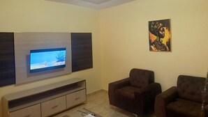 Un salon avec un téléviseur à écran plat fixé sur un meuble en bois, deux fauteuils marron, un tableau au mur et un luminaire au plafond.