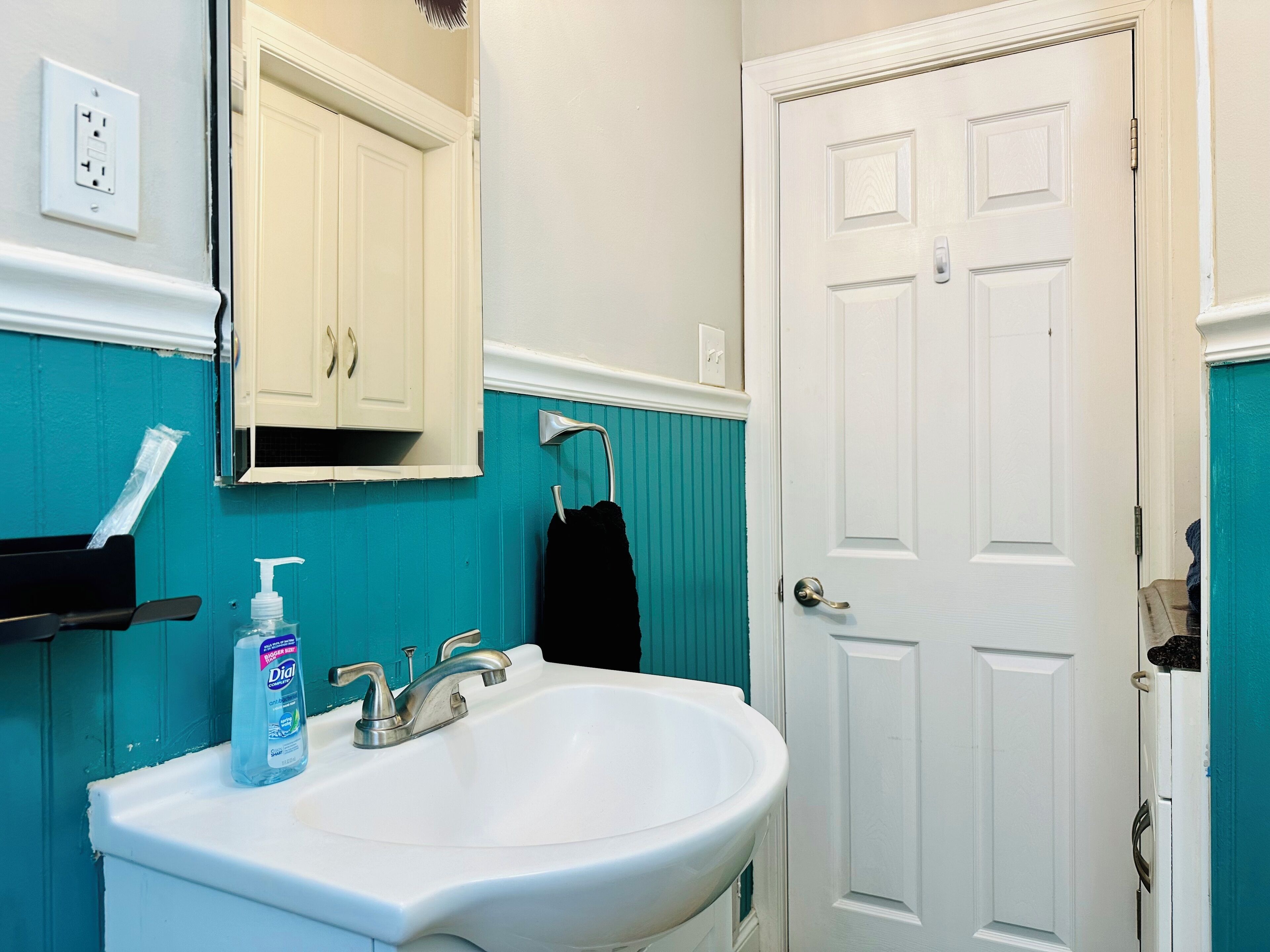 Un bagno con un lavandino bianco, uno specchio sopra, una parete verde acqua, una porta bianca e un asciugamano nero appeso a un gancio.