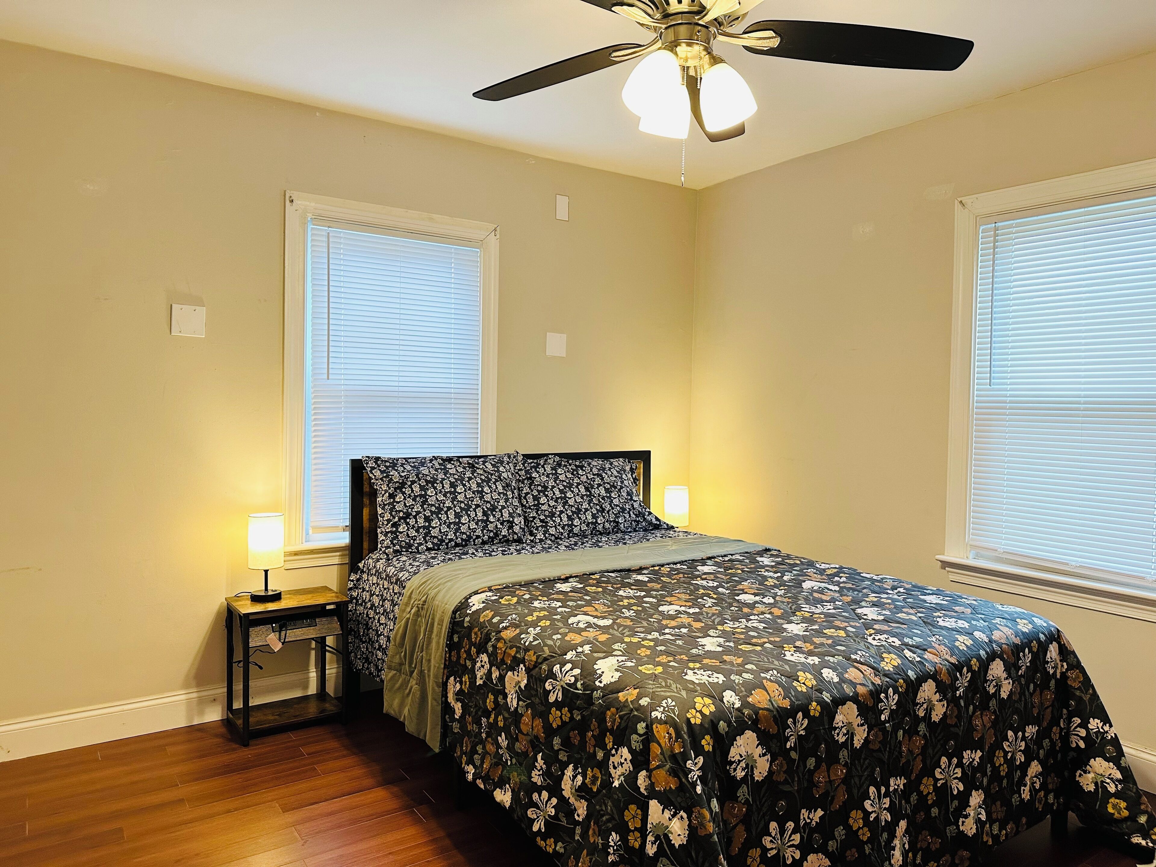 A bedroom with a bed, two bedside lamps, a ceiling fan, and two windows with blinds.