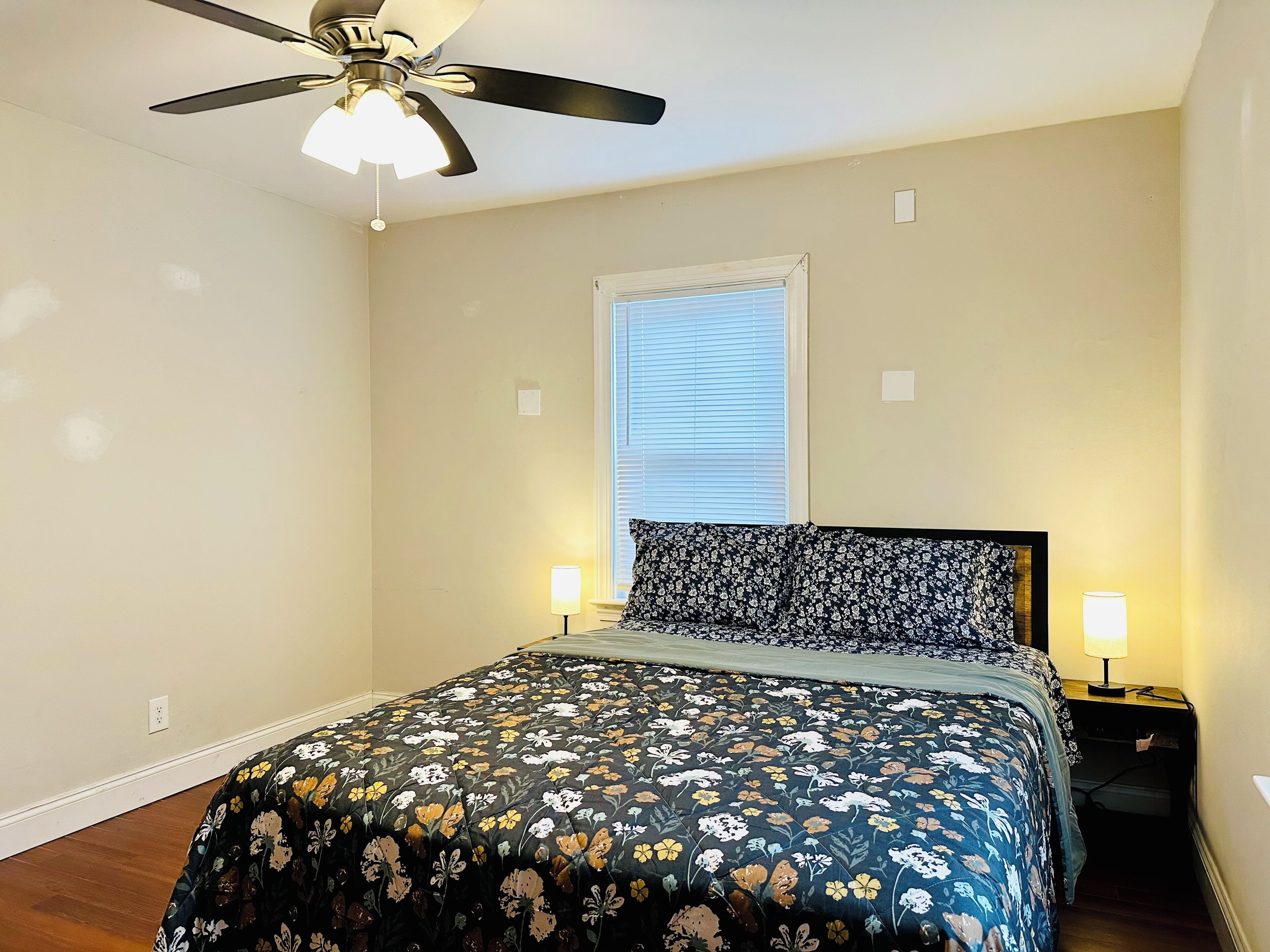 A bedroom with a bed, a ceiling fan, a window with blinds, and two bedside lamps.