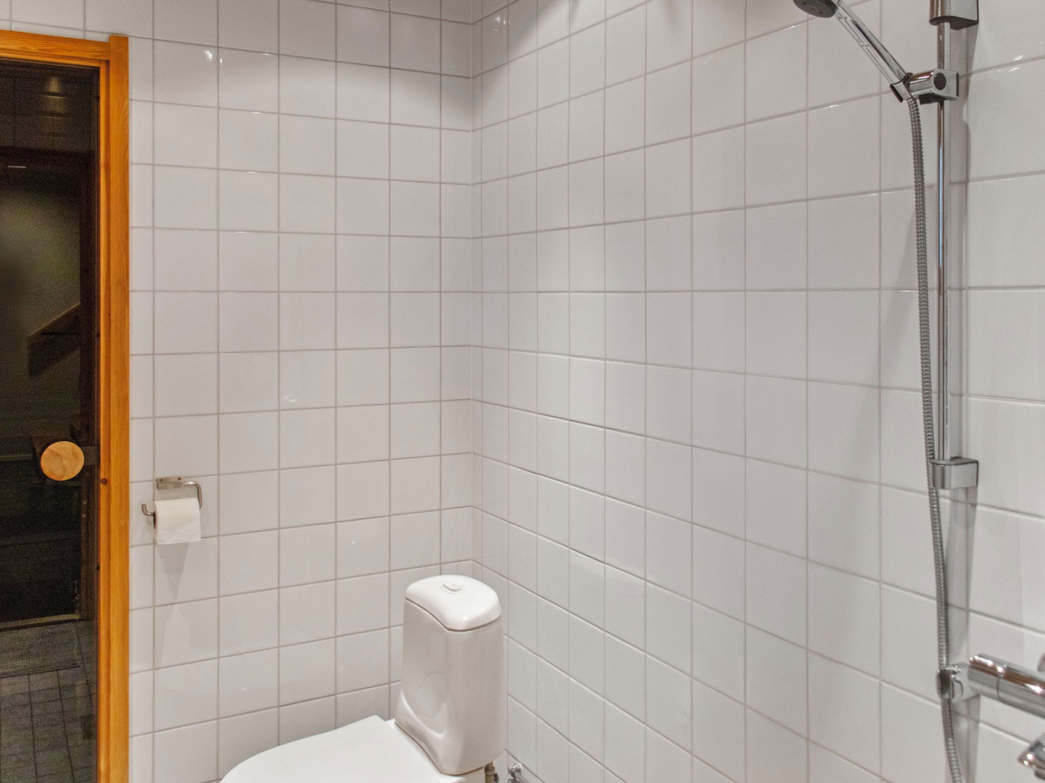 A bathroom with white tiled walls, a toilet, a shower with a handheld hose, and a door with a wooden frame.