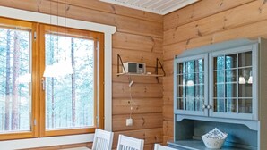 Interior de una cabaña de madera con mesa de comedor, sillas, zona de cocina con armarios y un amplio ventanal con vistas a árboles.
