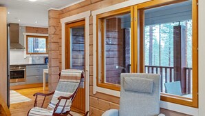 Interior de una cabaña con cocina, una mecedora, un sillón con cojines y ventanas amplias con vistas al bosque.