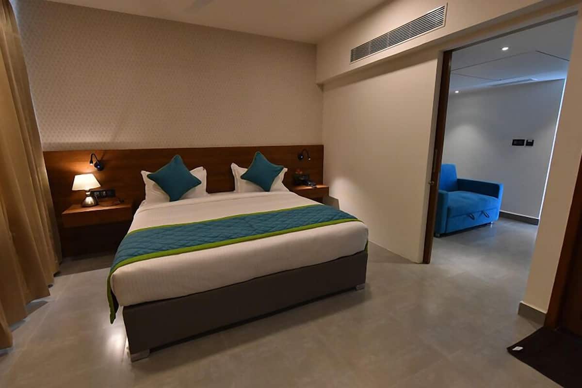 A hotel room with a large bed, a bedside table with a lamp, a nightstand, and a blue chair in the adjacent room.