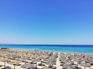 Une plage bordée de nombreux parasols en chaume et de chaises longues, un ciel bleu azur et l’océan en toile de fond.