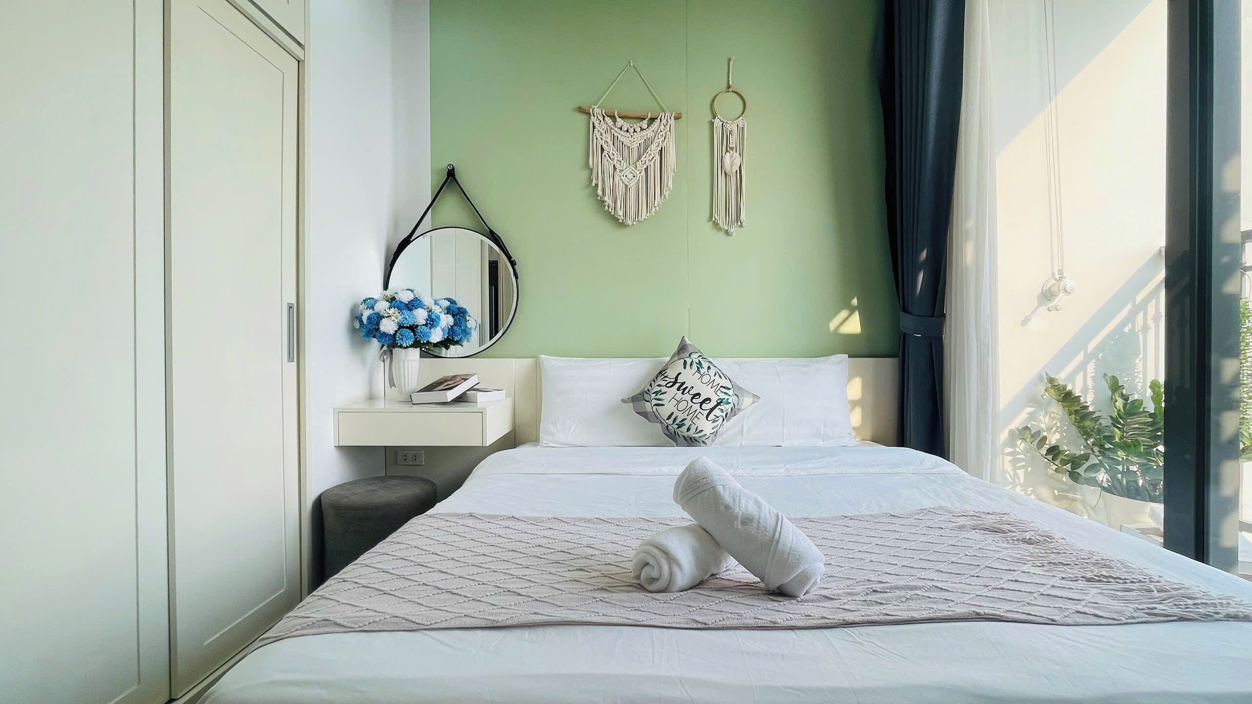 A neatly made bed with a rolled towel, a bedside table with a vase of flowers, a mirror, and macrame wall hangings.