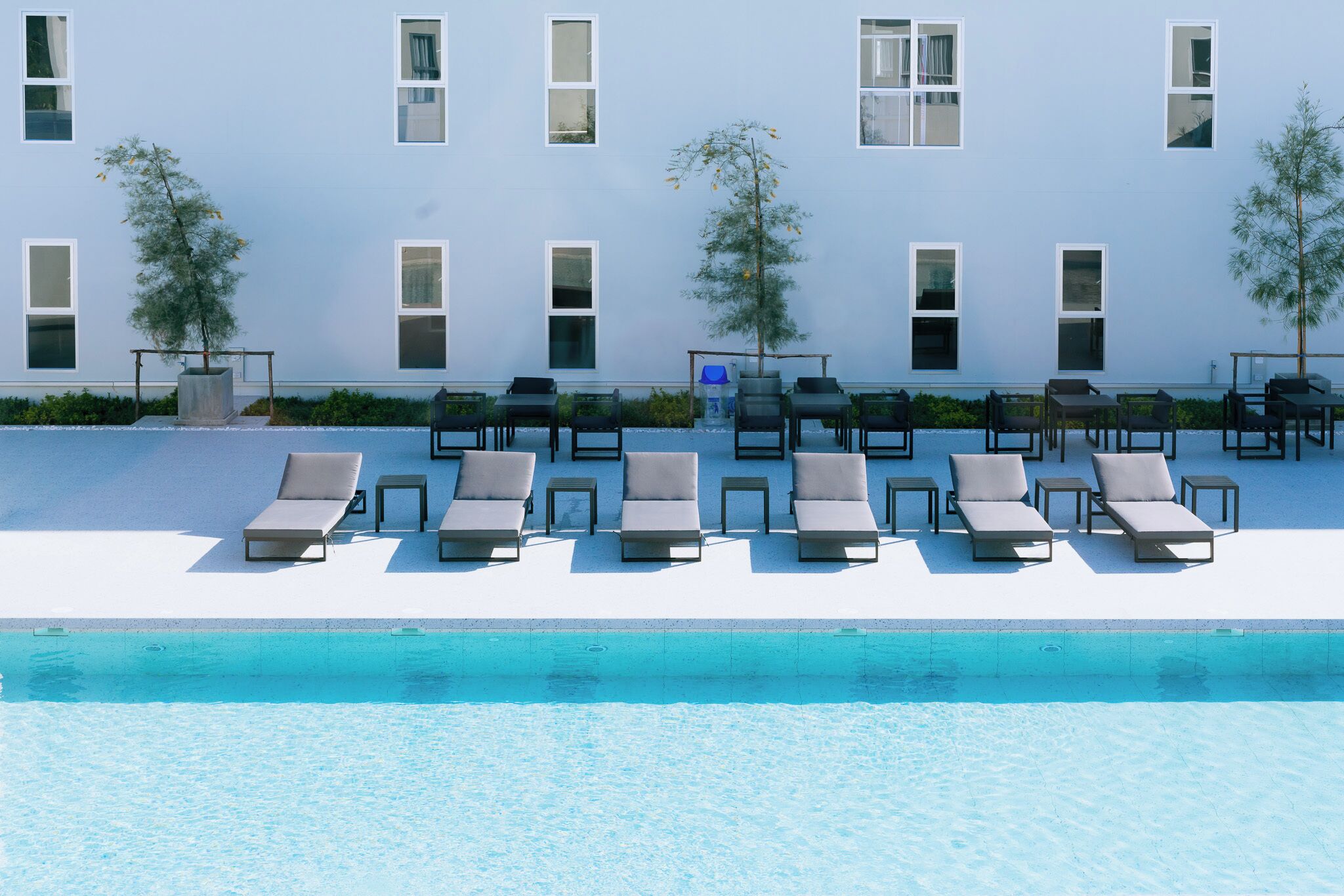 A poolside area with lounge chairs and tables, a clear pool, and a building with multiple windows.