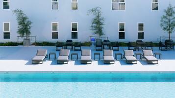 A poolside area with lounge chairs and tables, a clear pool, and a building with multiple windows.